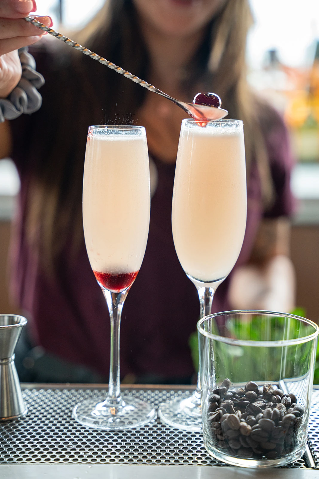 A close-up shot focuses on a bartender’s hand holding a long spoon with a cherry delicately placed at the tip, adding the finishing touch to a full glass of champagne. The glass sparkles with bubbles, and the bartender’s hand adds an elegant touch to the scene, creating a refined and thoughtful presentation.