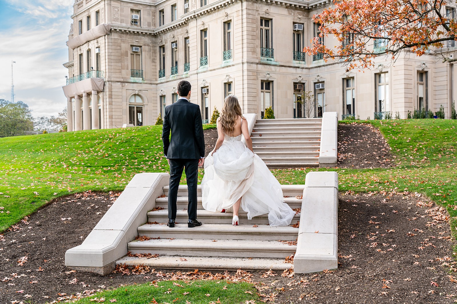 A back shot of a bride and groom walking up the steps of Monmouth College, facing the grand building. The bride delicately lifts her dress as she ascends, while the groom walks beside her. The blue sky with white clouds frames the scene, adding a sense of elegance and anticipation to the moment.