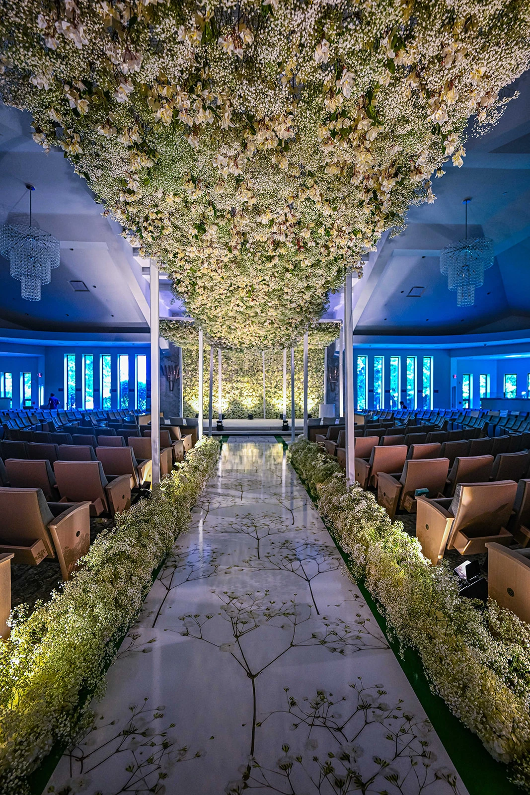 A beautifully arranged ceremony area glows under dimmed lighting, creating an intimate and romantic ambiance. Rows of chairs are neatly aligned, leading to a stunning aisle adorned with white flowers. The floral arrangements continue up a graceful archway and onto the ceremony stage, where the soft lighting enhances the elegant and serene atmosphere.