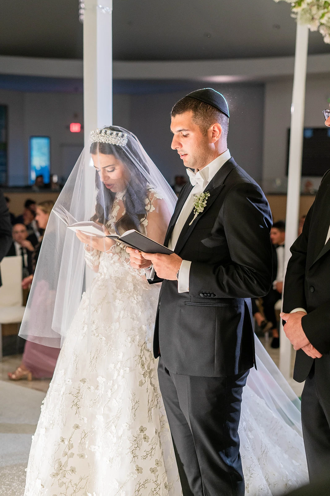 A Jewish bride and groom stand together under the huppah, deeply engaged in prayer and reading from their prayer books. Both hold their books in their hands, staring down with focus and reverence. The soft light and the sacred setting create an intimate and spiritual atmosphere as they share this meaningful moment in their ceremony.