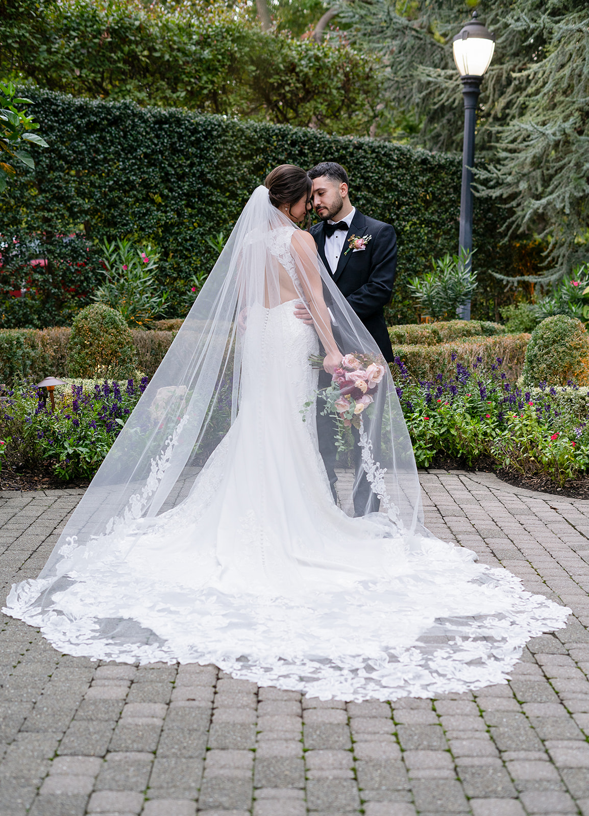 A bride and groom stand outside, face to face at a side angle, lost in a loving gaze. The groom gently rests his hand on the bride’s waist, holding her close, while her flowing train and delicate veil spread out beautifully in front of them. The soft natural light enhances the romance of the moment, capturing their deep connection and the elegance of their wedding attire.