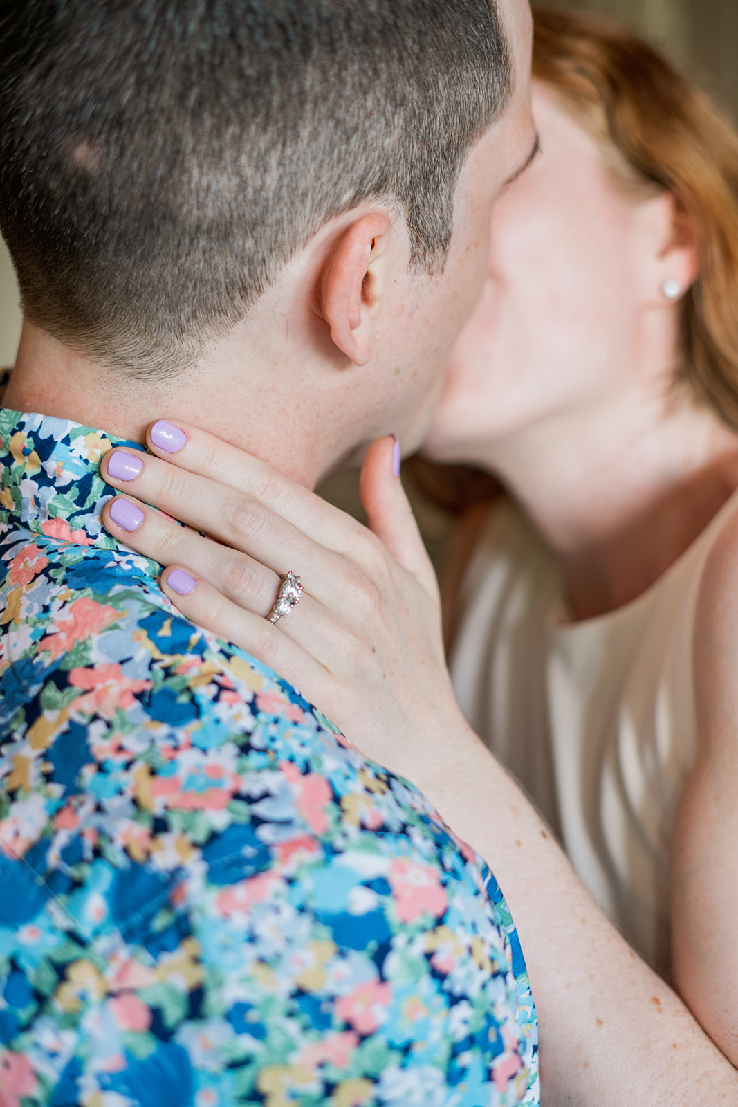 A close-up shot of a couple sharing a kiss, with the focus on the woman's hand gently resting on her partner's face, showcasing her sparkling engagement ring. The background is blurred, with soft lighting emphasizing the details of the ring and the intimacy of the moment. The couple’s blurred faces add to the romantic and dreamy atmosphere.