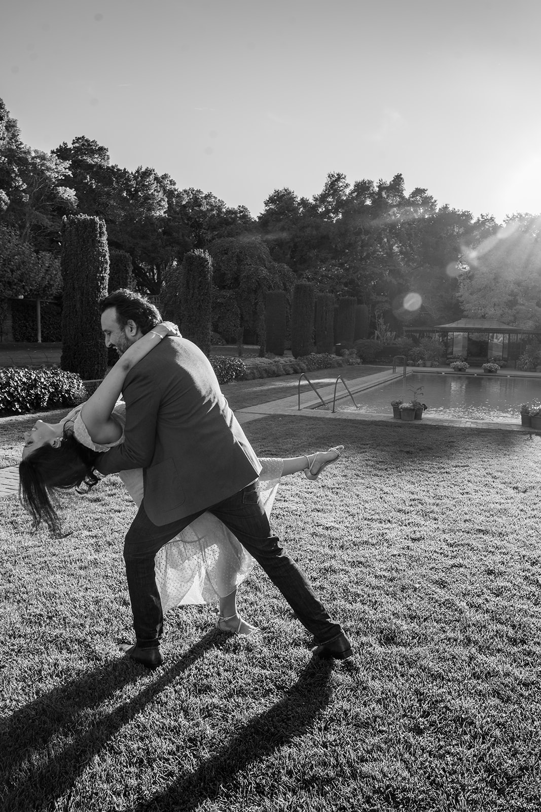 A black and white photo of a couple dancing in the gardens of Filoli in California, with the man gracefully dipping his fiancée as she smiles and laughs. A soft ray of sunshine cuts in from the side, adding depth and warmth to the moment. The surrounding lush greenery frames the couple, enhancing the romantic and timeless feel of the image.