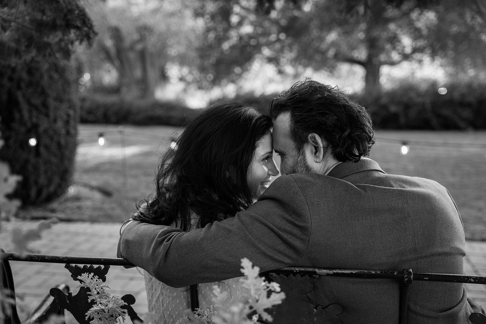 A black and white photo of a couple sitting closely on a bench, their foreheads gently touching. They are smiling into each other’s eyes, their expressions full of love and connection, creating an intimate and tender moment.