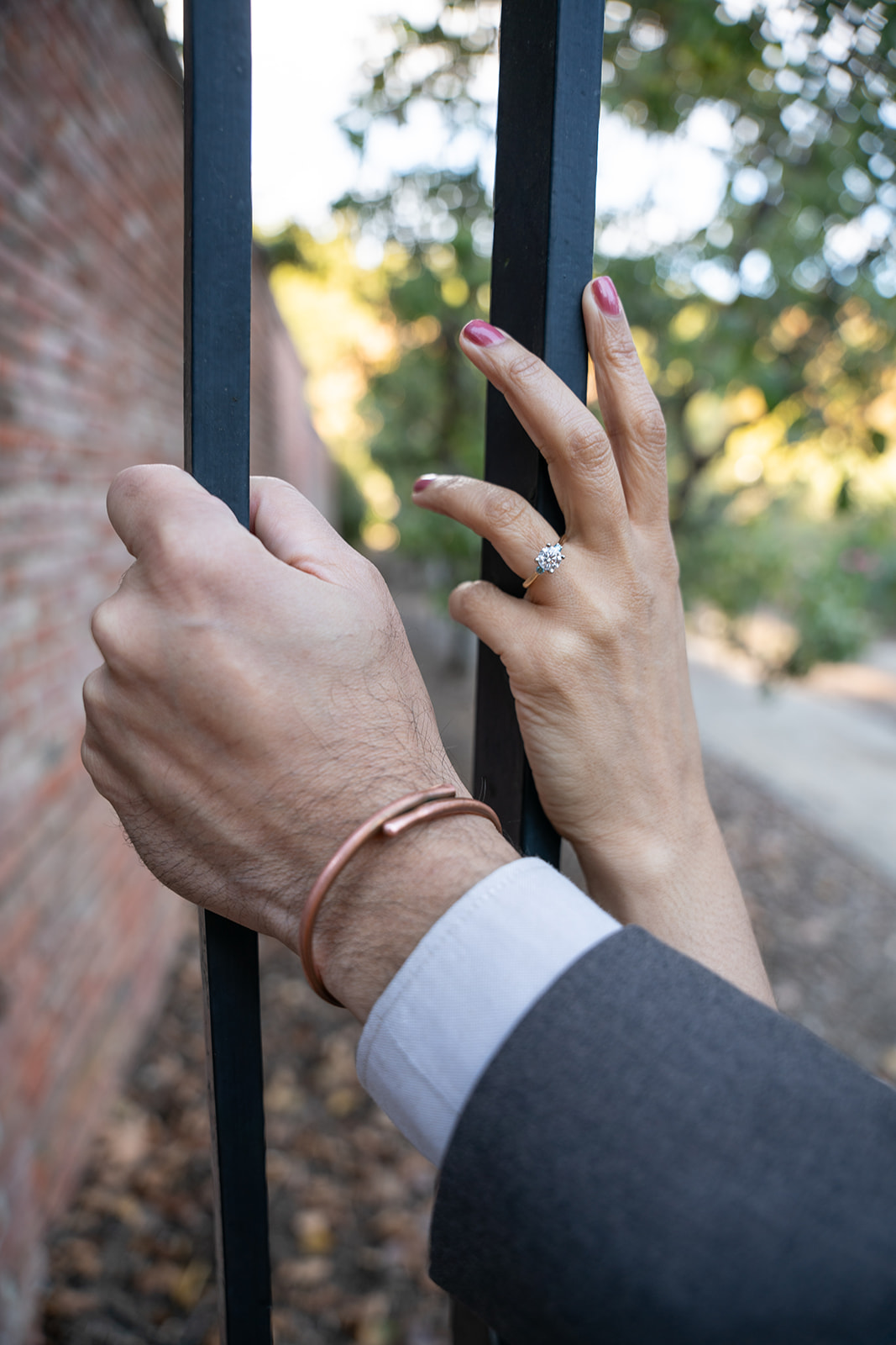 A close-up shot focuses on the couple’s hands as they each hold separate black bars on a gate, positioned next to each other. The image highlights the bride’s engagement ring, catching the light and sparkling with elegance. Their hands rest side by side, symbolizing their connection and the beginning of their journey together.