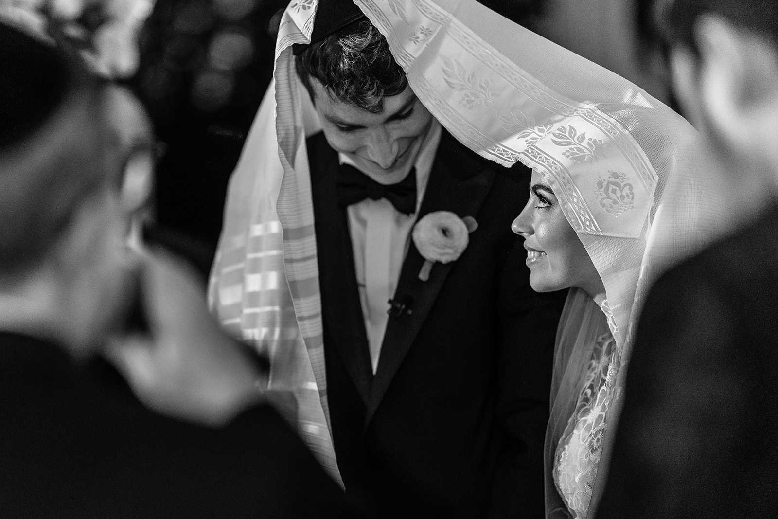 A bride and groom gaze at each other lovingly from beneath the tallit, their faces softly illuminated by the fabric’s gentle folds. They smile at each other, their expressions filled with warmth and devotion. The tallit drapes over them like a sacred canopy, symbolizing their union and shared faith.A bride and groom gaze at each other lovingly from beneath the tallit, their faces softly illuminated by the fabric’s gentle folds. They smile at each other, their expressions filled with warmth and devotion. The tallit drapes over them like a sacred canopy, symbolizing their union and shared faith.