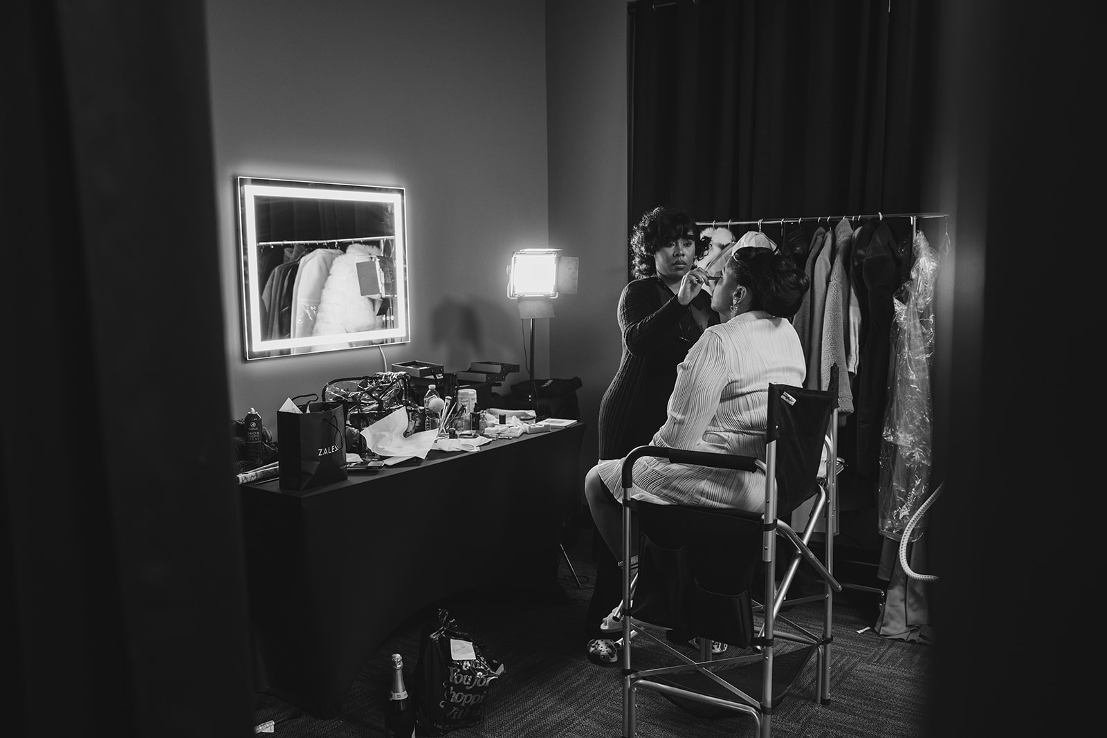 A black and white photo captures a makeup artist carefully applying makeup to a bride in a quiet dressing room as she prepares for her wedding day. The artist’s focused expression contrasts with the bride’s calm demeanor, her eyes gently closed. Soft natural light filters through the space, highlighting the delicate details of the moment, from the makeup brush’s subtle movement to the bride’s serene anticipation.