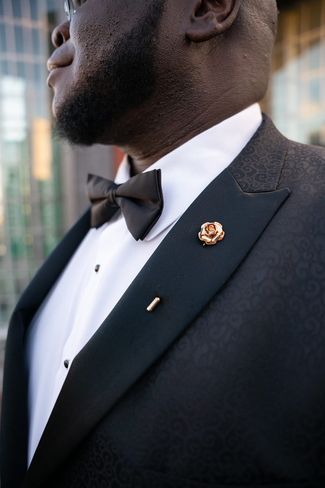 A close-up shot of a groom with the camera focused on his rose-shaped pin. The intricate details of the pin stand out against the texture of his suit, adding a touch of elegance. The blurred background subtly hints at the wedding setting, emphasizing the refined accessory and the groom’s polished look.