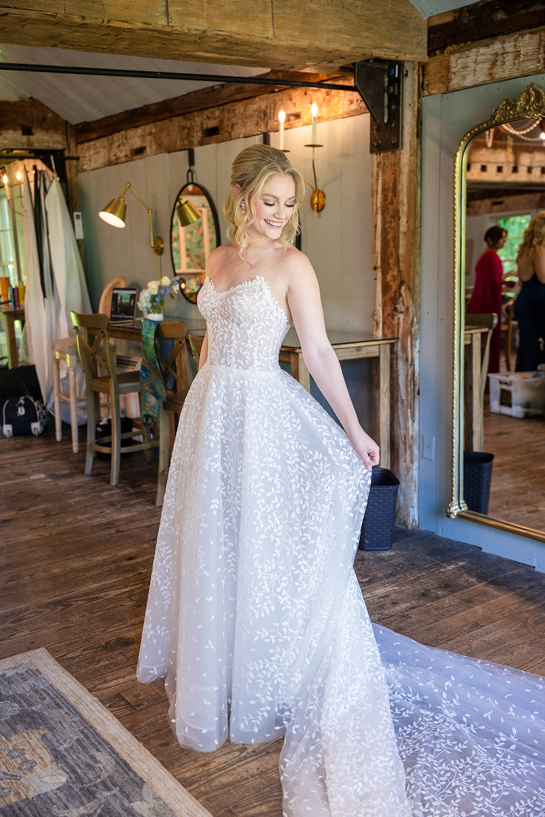 A bride smiles softly as she plays with her wedding dress, gently lifting the fabric while admiring the flowing train beneath her. The delicate details of the gown catch the light, adding to the sense of joy and anticipation in this intimate moment.
