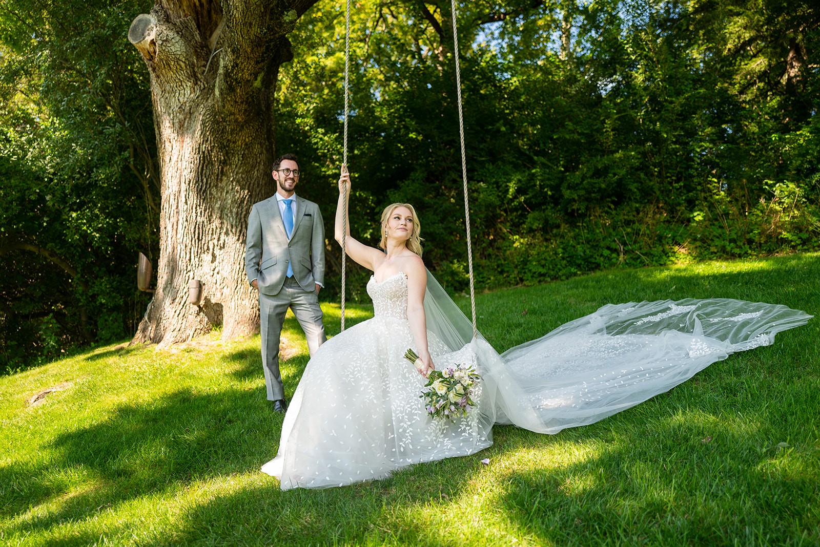 A bride in a strapless white gown with delicate floral appliqués sits gracefully on a rope swing, holding a bouquet of white and pastel flowers. Her long veil cascades onto the green grass, creating a flowing train behind her. She gazes upward with a serene expression. The groom, dressed in a light gray suit with a blue tie, stands nearby with a warm smile, his hands in his pockets. They are positioned under a large tree with textured bark, surrounded by lush greenery and dappled sunlight filtering through the leaves