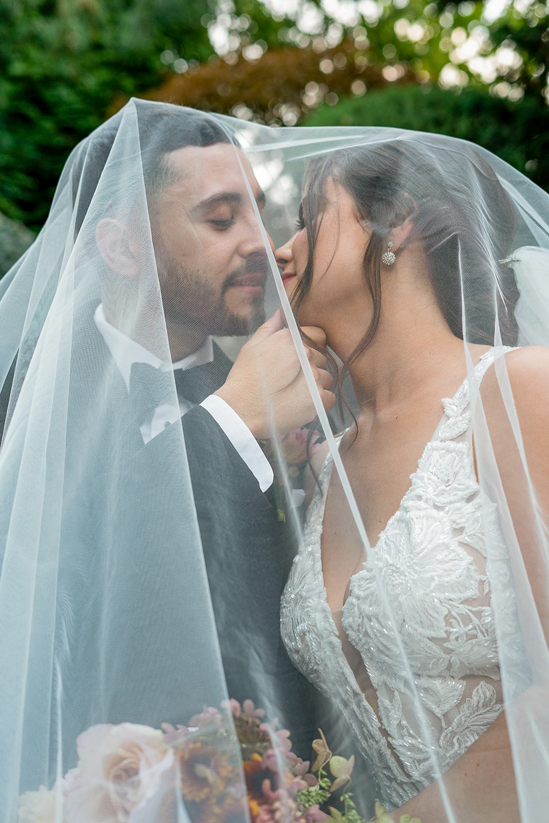 A couple sits face to face under the bride’s veil, their gazes fixed on each other’s lips with a sense of anticipation. The groom gently holds the tip of the bride’s chin, guiding her closer to him. The soft fabric of the veil creates a dreamy, intimate atmosphere as they share a quiet, tender moment before their kiss.