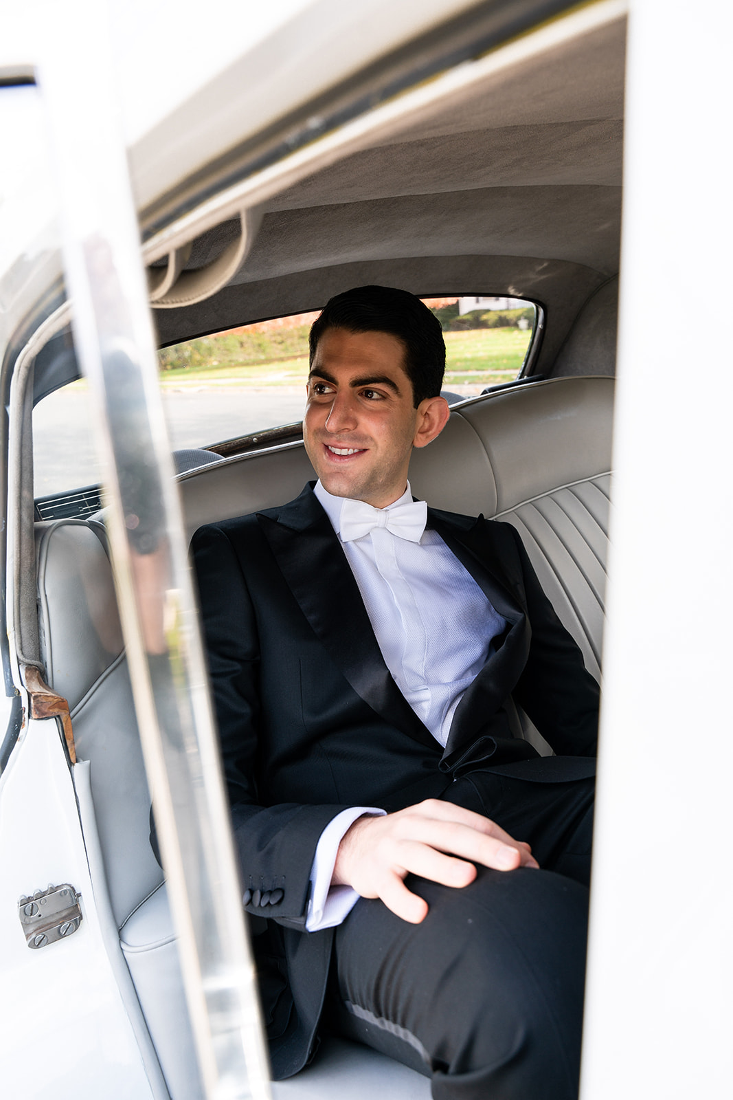 A groom sits inside a luxurious Rolls-Royce, smiling as he gazes sideways. The image is captured through the narrow crack of the open car door, adding a sense of depth and intimacy. Soft light highlights his joyful expression, while the elegant interior of the car subtly frames the moment.