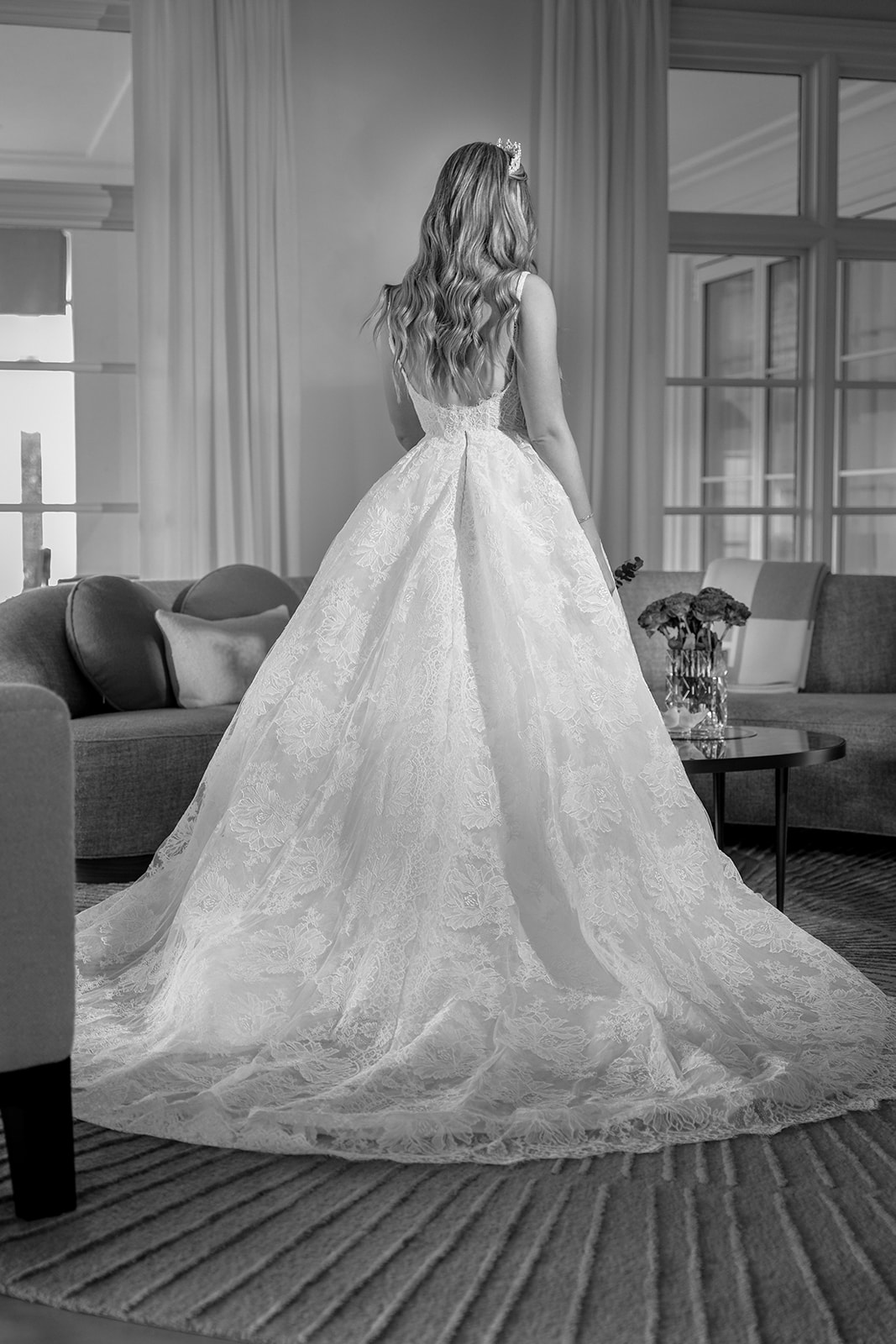 A black and white image of a bride standing in her living room, facing the window. Soft natural light outlines her silhouette, highlighting the delicate details of her gown and veil. Her posture conveys a quiet moment of reflection, with the contrast of shadows and light adding depth and emotion to the scene.
