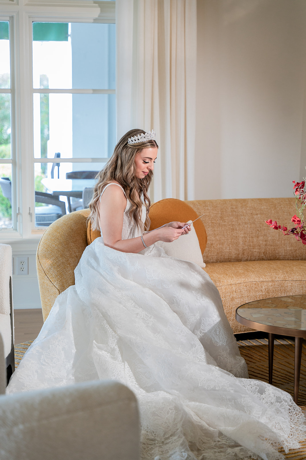 A bride sits gracefully in her living room, immersed in reading a heartfelt love note from her soon-to-be husband. A soft smile spreads across her face as she takes in his words, her eyes filled with emotion. Natural light filters through the room, casting a warm glow on her wedding dress, creating a serene and intimate moment before the ceremony