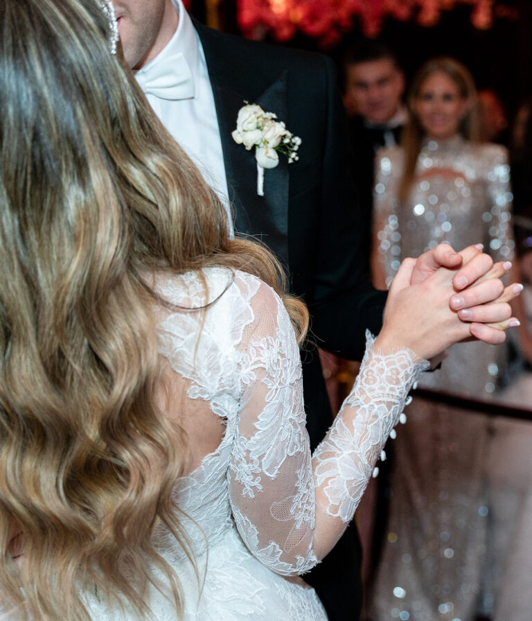 A close-up shot of a newly married couple’s hands gently intertwined as they share their first dance. The bride’s delicate wedding band glimmers under the soft, ambient lighting, while the groom’s hand firmly yet tenderly holds hers. The blurred background hints at the warm glow of the reception, evoking a sense of intimacy and romance.