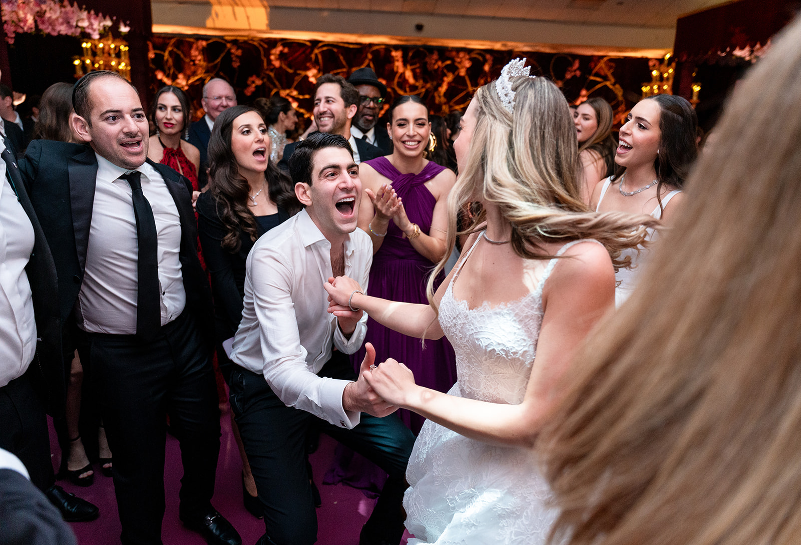 A joyful couple dances hand in hand on the dance floor, surrounded by cheering guests in the background. Their movements radiate love and excitement as the warm glow of the venue lights highlights the moment. The guests' smiles and raised hands add to the celebratory energy, capturing the essence of a joyous wedding reception.