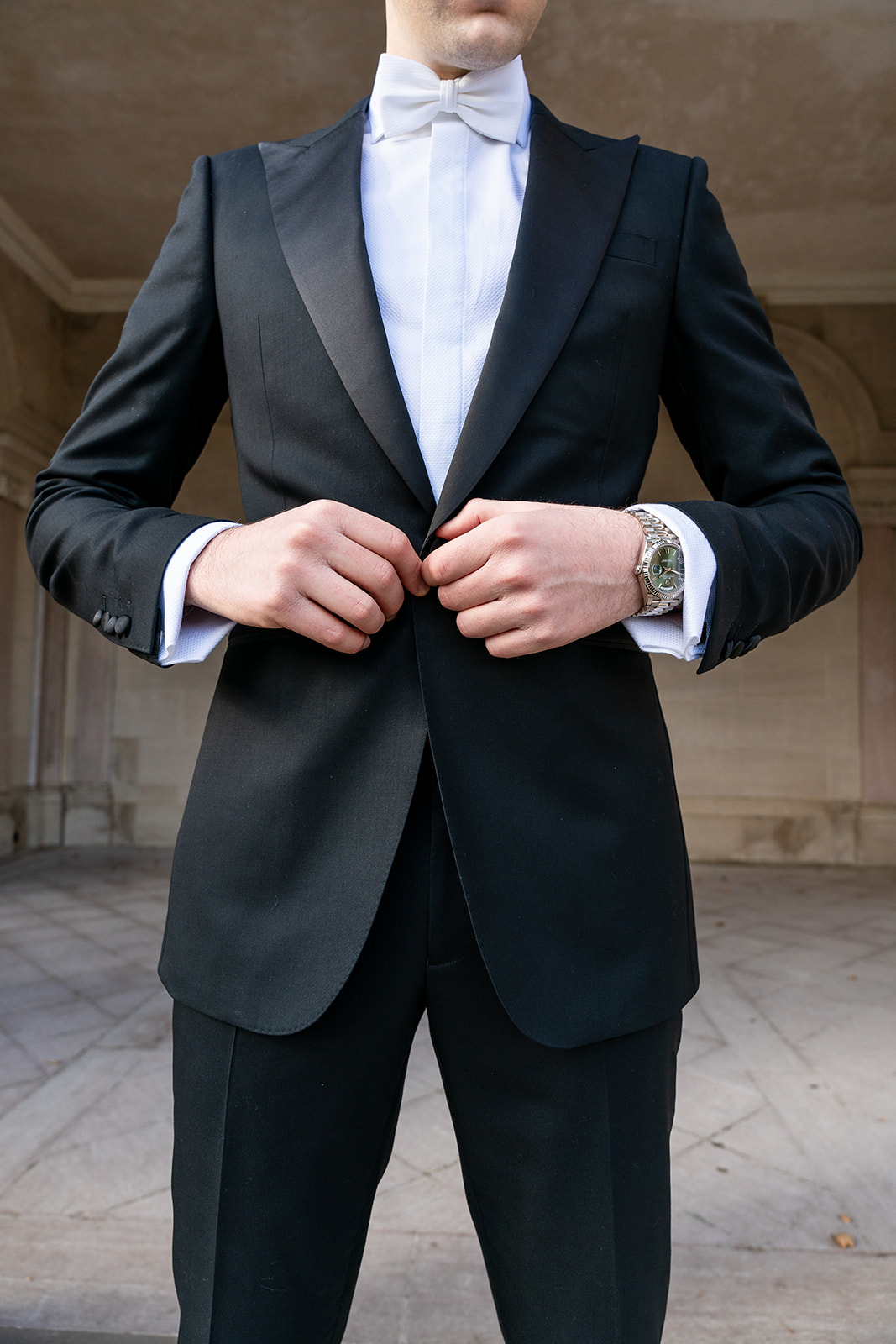 A close-up shot captures a groom buttoning his blazer, his hands carefully fastening the button with precision. The rich texture of the fabric and subtle details of the suit are in sharp focus, highlighting the elegance of his attire. The image exudes a sense of anticipation and refinement as he prepares for his special moment.