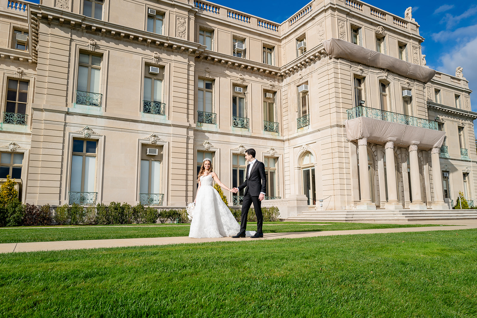 A newlywed couple walks hand in hand on a lush green lawn in front of Monmouth University, an elegant, historic building with intricate architectural details and soft beige tones. The bride wears a flowing white wedding gown with a delicate tiara, while the groom looks dapper in a classic black suit. The scene is bathed in natural sunlight, enhancing the romantic and timeless feel of the moment.