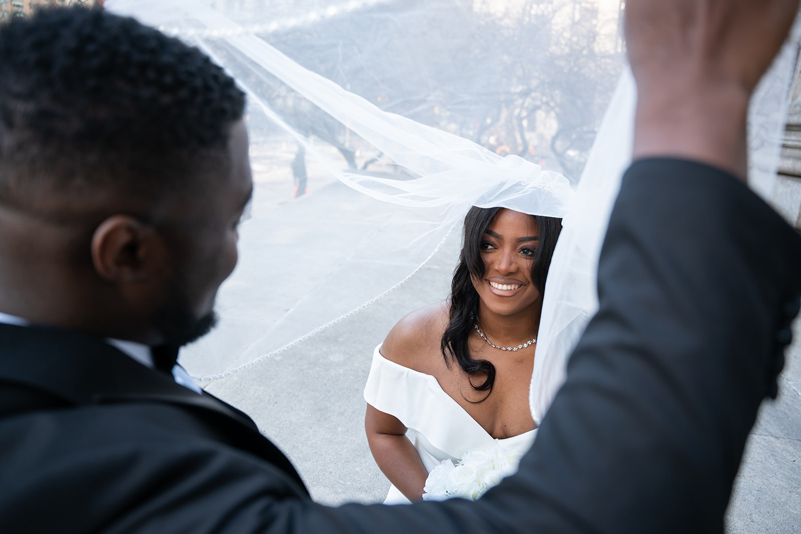 A groom playfully peeks under his wife's veil outdoors as she tilts her head back, smiling up at him. Soft natural light highlights their joyful expressions, with the delicate veil adding a dreamy, romantic touch to the moment. The background is softly blurred, keeping the focus on their love and connection.