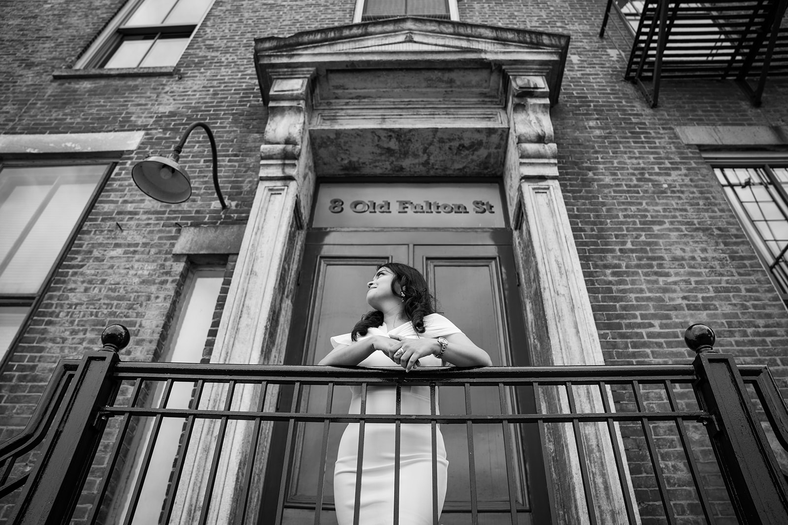 A black and white image of a woman in a white dress, leaning over a gate and looking away. The scene is set under an old Fulton Street brownstone, with the architectural details adding depth to the moment. The woman's posture and distant gaze create a sense of elegance and quiet contemplation.