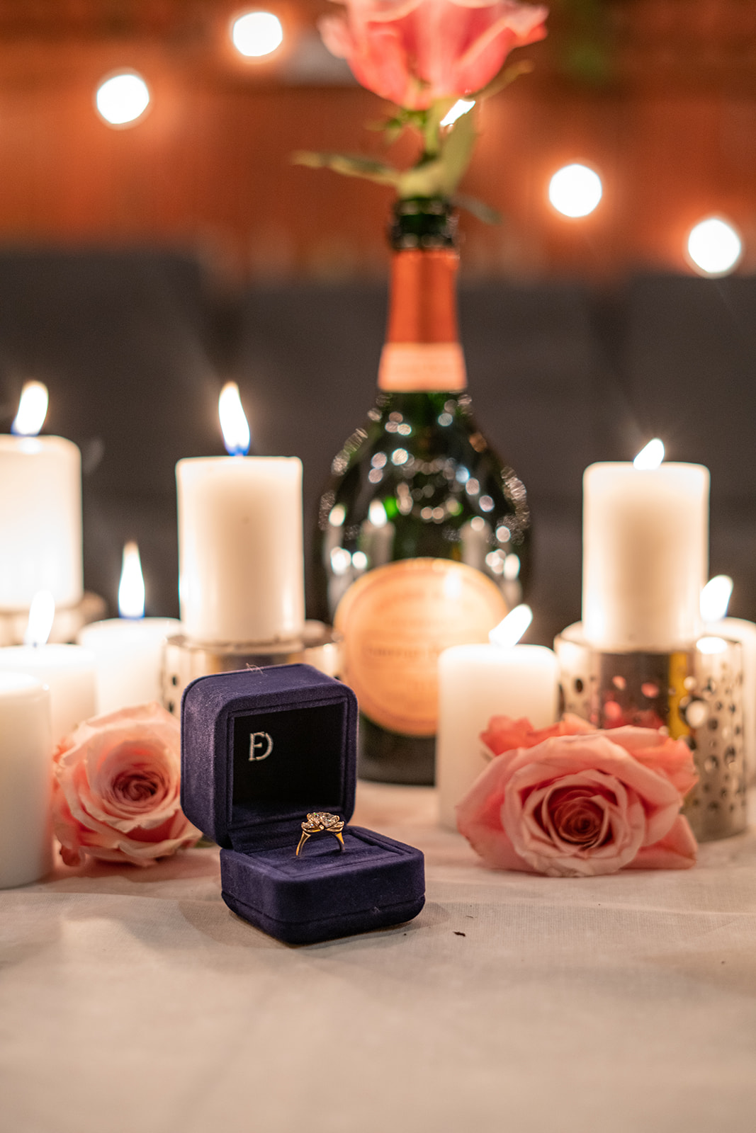 A nighttime scene with a table illuminated by glowing candles. In front of an empty Laurent Perrier champagne bottle, an open engagement ring box displays a sparkling ring. Two pink roses rest behind the box, while another rose sits inside the bottle, adding a romantic and elegant touch to the setting.