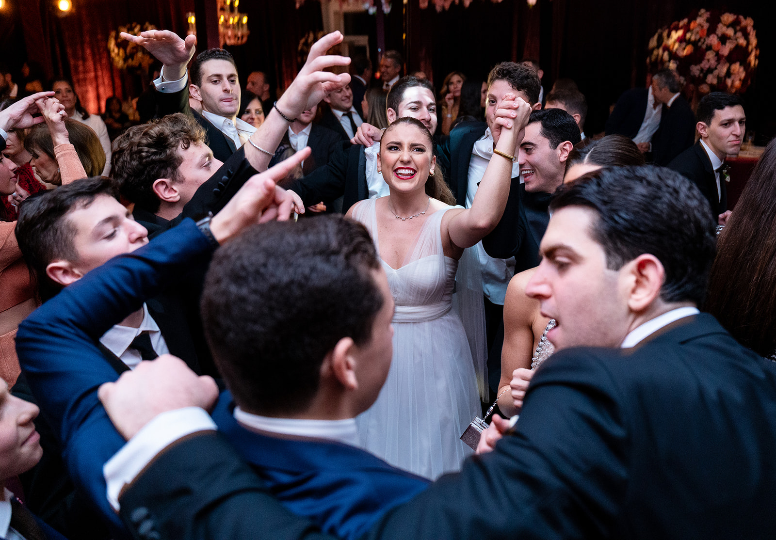 A joyful wedding reception scene with guests dancing in a circle, their hands linked as they sway. The camera focuses through the open space in the circle, capturing a young woman at the center, singing with passion and dancing with one hand raised. Warm ambient lighting enhances the celebratory atmosphere, with blurred movement of the guests adding to the sense of energy and excitement.