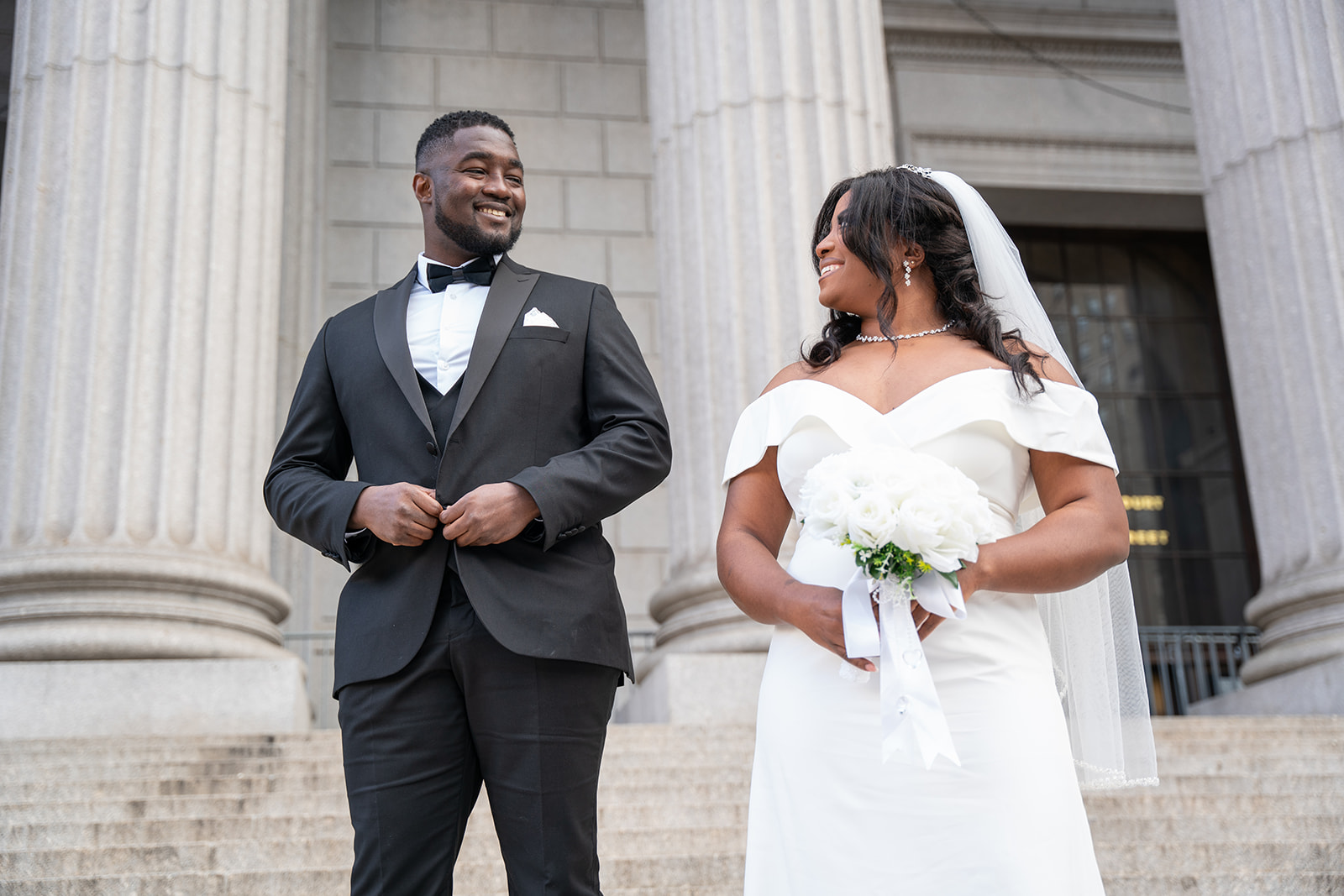 A beautiful Black couple stands side by side on the steps of City Hall, smiling warmly at each other as they prepare to elope. Dressed elegantly, they share a joyful and intimate moment, with the grand architecture of the building serving as a timeless backdrop.