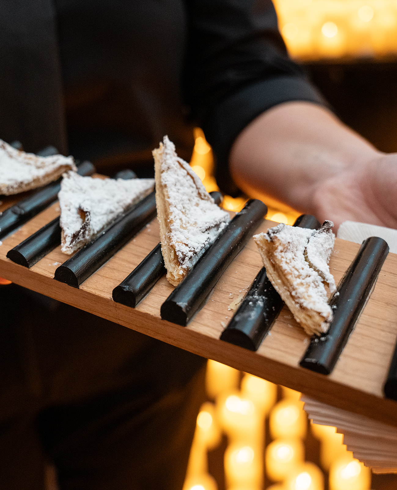 A close-up of a delicate apple dessert dusted with powdered sugar, held by a caterer’s hand above softly glowing candles. The warm candlelight highlights the dessert’s texture, creating a cozy and inviting atmosphere.