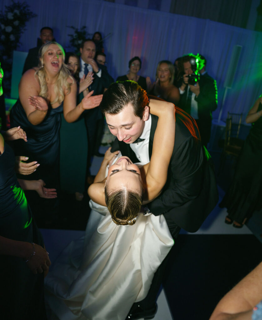 white groom in his black tuxedo dips his white bride in her white dress in the middle of the black and white checkered dance floor at their wedding reception and they both are smiling at one another while their friends and family watch and cheer them on