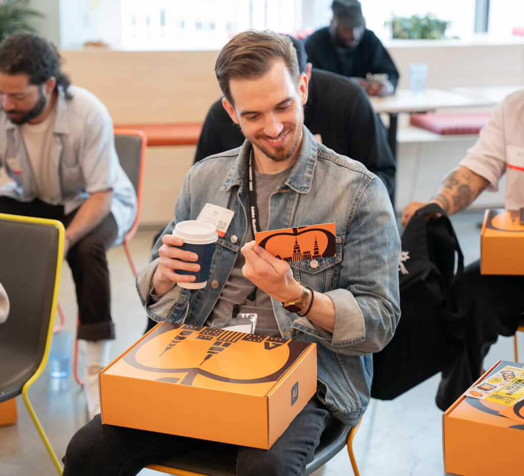 Adobe Creative attendee checking out his partnership box while holding a cup of coffee smiling at the thank you letter given to him by Adobe Leaders which came in a bright orange box that sit on his lap while he wears a blue jean jacket