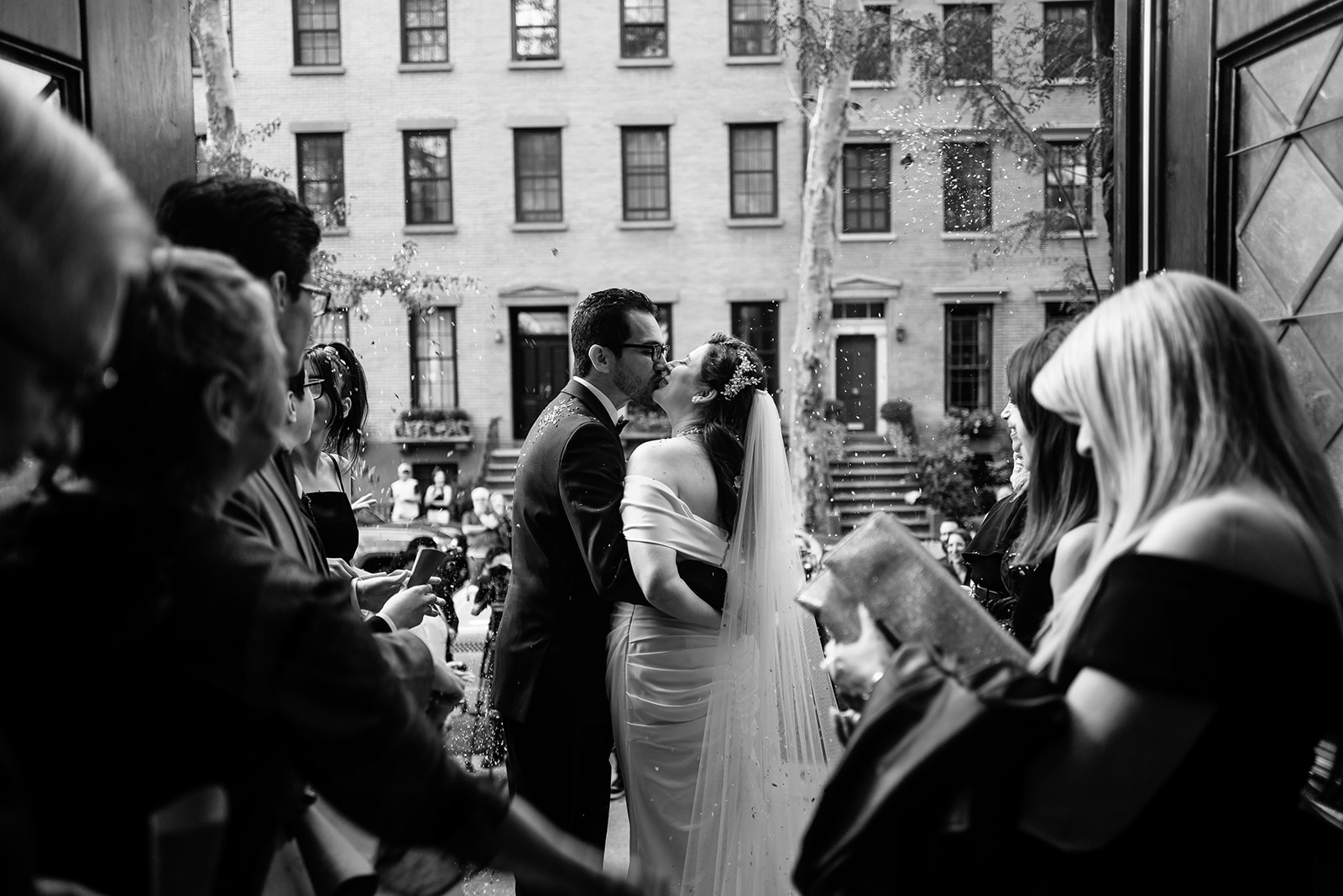 Black and white photo of a beautiful spanish couple kissing in front of a church with their family and friends watching throwing rice at them while the church doors are open in Brooklyn Heights