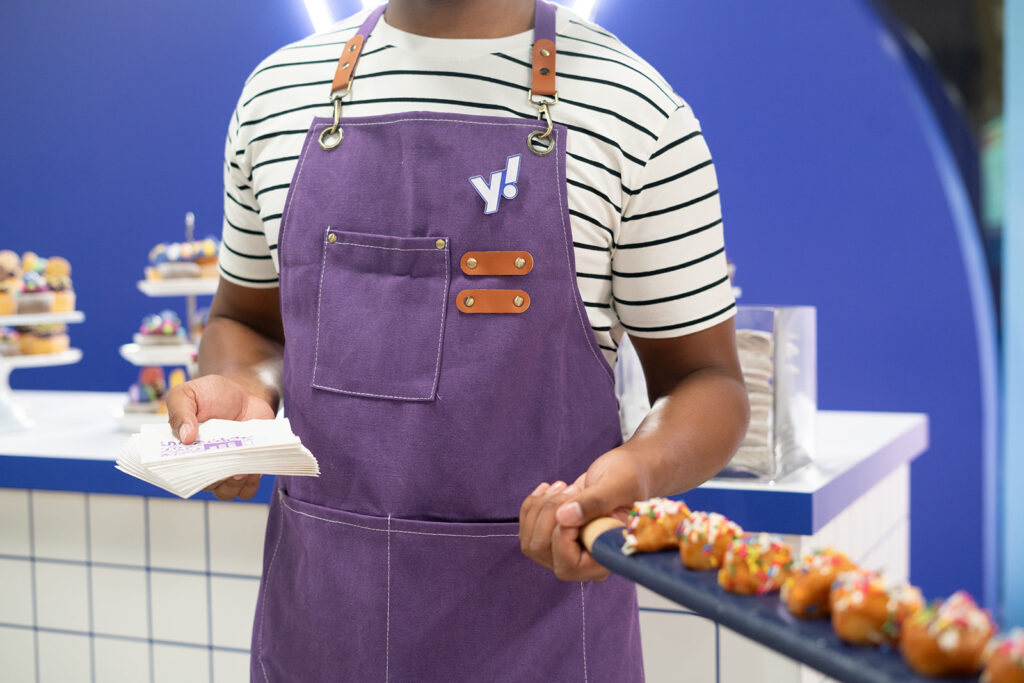 someone holding a long platter with tiny glazed donuts with sprinkles holding a handful of napkins with the Yahoo symbol showing on their purple apron wearing a white and blue striped shirt from the neck down
